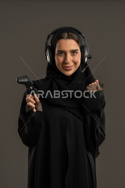 ايماءات الفوز والانتصار ، الألعاب الالكترونية والأجهزة الحديثة ، العالم الرقمي الترفيهي ، اللعب و التسلية بالعاب الفيديو ، بورترية لامراة عربية خليجية إماراتية ترتدي العباءة السوداء والحجاب ، تمسك بيدها عصا التحكم بتركيز , سيدة سعودية تنظر الى الكاميرا بتعابير السعادة ، الاستمتاع بتجربة ترفيهية مثيرة ،خلفية سوداء .