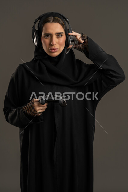 انشطة شبابية ممتعة ، الألعاب الالكترونية والأجهزة الحديثة ، العالم الرقمي الترفيهي ، اللعب و التسلية بالعاب الفيديو ،بورترية لامراة عربية خليجية إماراتية ترتدي العباءة السوداء والحجاب ، تمسك عصا التحكم بتركيز , سيدة سعودية تنظر الى الكاميرا وتمسك سماعة الرأس بيدها ،الاستمتاع بتجربة ترفيهية مثيرة ،خلفية سوداء .