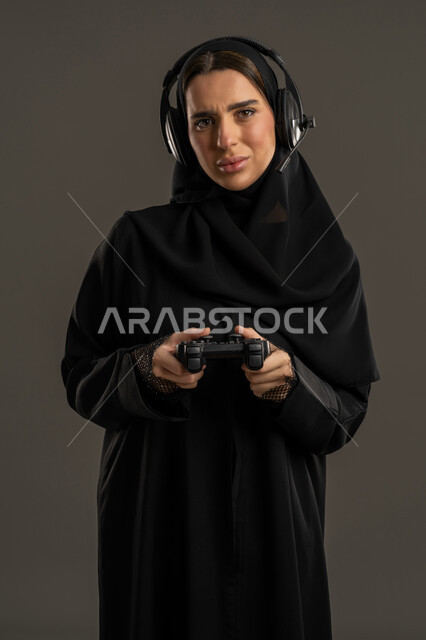 النظر الى الكاميرا بايماءات الغضب ، الألعاب الالكترونية والأجهزة الحديثة ، العالم الرقمي الترفيهي ، المتعة والتسلية بالعاب الفيديو ، بورترية لامراة عربية خليجية إماراتية ترتدي العباءة السوداء والحجاب وسماعة الرأس ، تمسك بيدها عصا التحكم بتركيز , سيدة سعودية تستمتع بتجربة ترفيهية ممتعة ومثيرة ،خلفية سوداء .