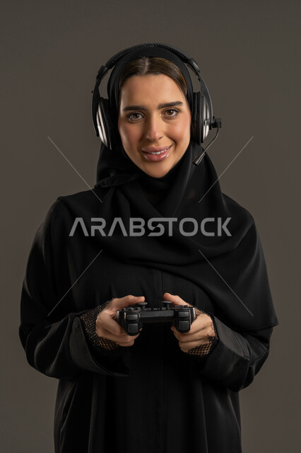 الاستماع الى الموسيقي عبر سماعات الرأس ، الحماس بالعالم الرقمي المتطور، الألعاب والأجهزة الالكترونية الحديثة ، اللعب و التسلية بالعاب الفيديو ، بورترية لامراة عربية خليجية إماراتية ترتدي العباءة السوداء والحجاب ، تمسك بيدها عصا التحكم بتركيز واندماج , سيدة سعودية تستمتع بتجربة ترفيهية مثيرة ،خلفية سوداء .