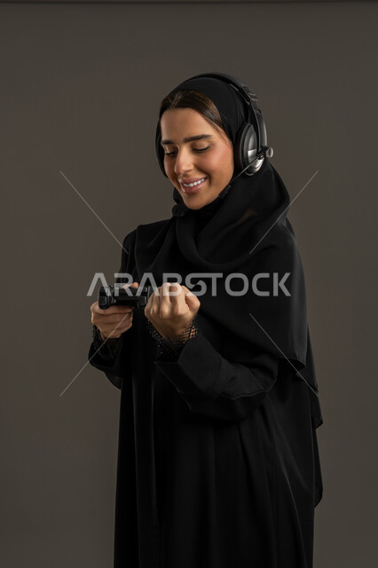 المتعة والتسلية بالعاب الفيديو, العالم الرقمي الترفيهي، بورترية لامراة عربية خليجية إماراتية ترتدي العباءة السوداء والحجاب وسماعة الرأس ، تمسك بيدها عصا التحكم بتركيز واندماج, سيدة سعودية تقبض يدها بايماءة الفوز والانتصار ، الاستمتاع بتجربة ترفيهية ممتعة ومثيرة في الألعاب الالكترونية والأجهزة الحديثة ،خلفية سوداء .