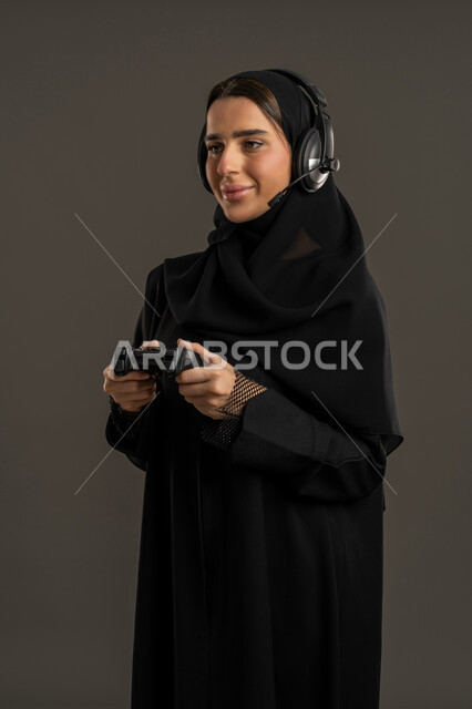 ايماءات الاندماج والتفاعل أثناء اللعب ، الاستمتاع والتسلية فى العطلة ، بورتريه لامرأة خليجية سعودية تضع سماعة الرأس الصوتية، تمسك بيدها عصا التحكم عن بعد ، سيدة سعودية تلعب بألعاب الفيديو الترفيهية ، إستخدام أجهزة تقنية حديثة ، خلفية سوداء .