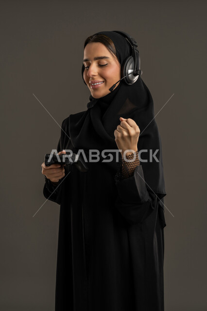 الاستمتاع والتسلية فى العطلة  ، إستخدام أجهزة تقنية حديثة ، بورتريه لامرأة خليجية سعودية تضع سماعة الرأس الصوتية، تمسك بيدها عصا التحكم عن بعد ، سيدة سعودية تلعب بألعاب الفيديو الترفيهية، ترفع قبضة اليد لاعلى بايماءات الفوز ، قضاء أوقات ممتعة ، خلفية سوداء .