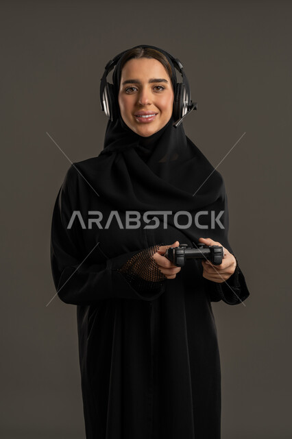 النظر الى الكاميرا بايماءات السعادة ، الاستمتاع والتسلية فى العطلة  ، إستخدام أجهزة تقنية حديثة ، بورتريه لامرأة خليجية سعودية تضع سماعة الرأس الصوتية، تمسك بيدها عصا التحكم عن بعد ، سيدة سعودية تلعب بألعاب الفيديو الترفيهية، خلفية سوداء .