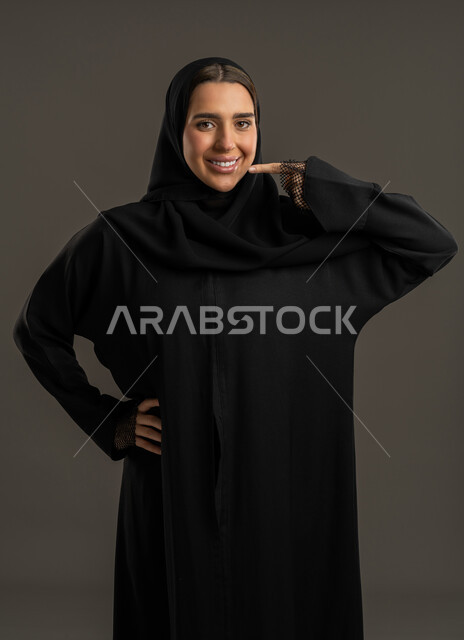 مفهوم الجمال والاهتمام بالمظهر الخارجي، بورترية مقرب لامرأة عربية خليجية اماراتية ترتدي العباءة  السوداء والحجاب ، تشير بإصبعها إلى فمها ، سيدة سعودية تنظر الى الكاميرا بايماءات السعادة ، خلفية سوداء.