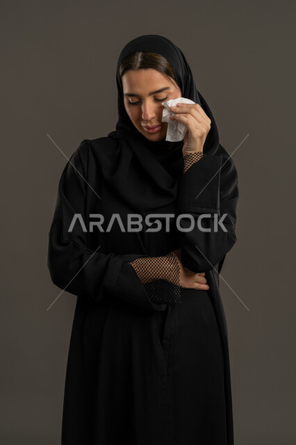 الوقوف بتعابير الخيبة واليأس, الشعور بالاكتئاب, بورتريه مقرب لامرأة عربية خليجية إماراتية ترتدي العباءة السوداء والحجاب تضع منديل على وجهها بحزن, سيدة سعودية تقف بإيماءات الهم والفقدان , خلفية سوداء .