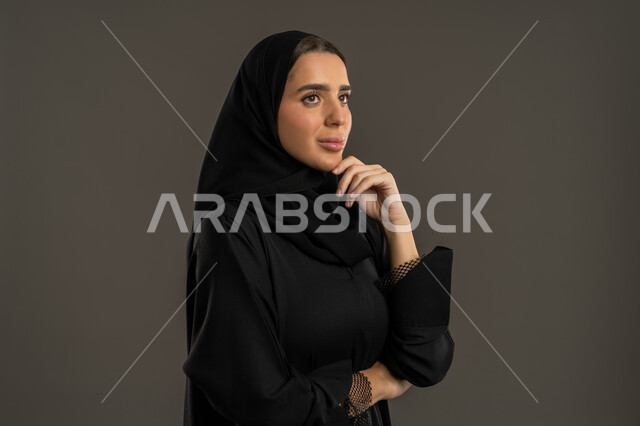إيماءات التأمل والتمعن ، بورتريه من الجانب مقرب لسيدة عربية خليجية اماراتية ترتدي العباءة السوداء والحجاب ، تنظر الى شئ ما بتعابير الاندماج و الحيرة ، سيدة سعودية  تضع يدها على ذقنها  لإيجاد الحلول لمشكلة ما ، خلفية سوداء .