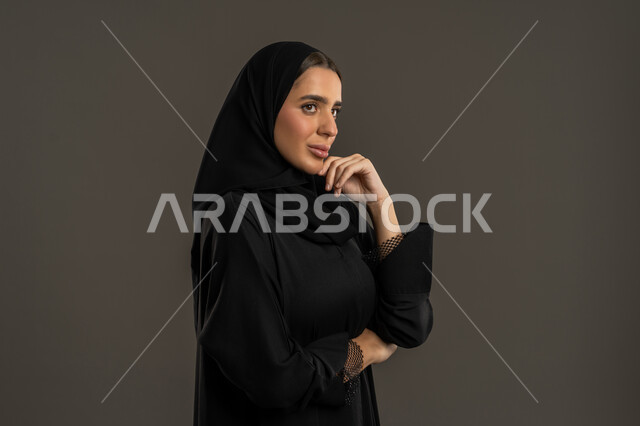 الشعور بالملل و شرود الذهن ، انتظار شيء ما ، بورتريه من الجانب مقرب لسيدة عربية خليجية اماراتية ترتدي العباءة السوداء والحجاب ، تنظر الى شئ ما بتعابير التركيز ، سيدة سعودية تضع يدها على ذقنها بإيماءات التمعن فى حل مشكلة ، خلفية سوداء .