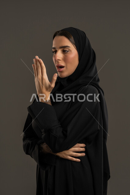 الشعور بالدهشة لحدوث أمر ما ،  بورتريه مقرب من الجانب لإمرأة عربية خليجية اماراتية ترتدي العباءة السوداء والحجاب ، تنظر الى يدها بايماءات الذهول والاستغراب ، سيدة سعودية تتعجب من شئ ما ، خلفية سوداء .