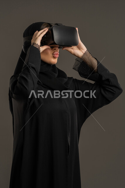 الاستمتاع بمشاهدة أفلام الواقع الافتراضي ، إيماءات التفاعل والاندماج مع العناصر الهولوجرامية ، دمج المتعة والتسلية مع التنقيات الحديثة ، بورتريه لامرأة عربية خليجية اماراتية ترتدي العباءة السوداء والحجاب ونظارة الواقع الافتراضى، سيدة سعودية تمسك بيدها نظارة الفي آر ، وسائل ترفيهية فى وقت الفراغ ، خلفية سوداء .