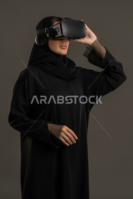 Using modern and advanced technical devices, combining the imaginary world with fun and entertainment. Gestures of interaction and integration with holographic elements. Entertainment during leisure time. Simulating the virtual world. A portrait of an Arab Gulf Emirati woman wearing a black abaya, hijab, and virtual reality (VR) glasses. A Saudi woman watching her favorite movies and TV series. A black background.