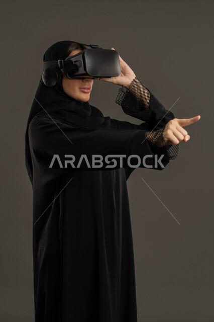 Combining the imaginary world with fun and entertainment. Gestures of interaction and integration with holographic elements. Entertainment during leisure time. Simulating the virtual world. A portrait of an Arab Gulf Emirati woman wearing a black abaya, hijab, and virtual reality (VR) glasses. A Saudi woman pointing her index finger at something. A technological device and modern technology. A black background.