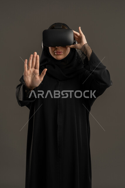Gestures of interaction and integration with holographic elements. Entertainment during leisure time. Simulation of the virtual world. Integrating fun and entertainment with the virtual world. A portrait of an Arab Gulf Emirati woman wearing a black abaya, hijab, and virtual reality (VR) glasses. A Saudi woman raising her hand to touch something. A technological device and modern technology. A black background.