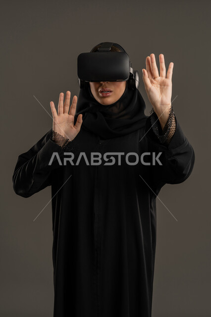 وسائل ترفيهية فى وقت الفراغ ، إستخدام حاسة اللمس لملامسة عناصر هولوجرامية ، محاكاة العالم الافتراضى ، دمج المتعة والتسلية مع العالم الافتراضي، بورتريه لامرأة عربية خليجية اماراتية ترتدي عباءة السوداء والحجاب ونظارة الواقع الافتراضى ( في آر )، سيدة سعودية ترفع كفة يدها لملامسة شئ ما ، جهاز تكنولوجي وتقنية حديثة ، خلفية سوداء .