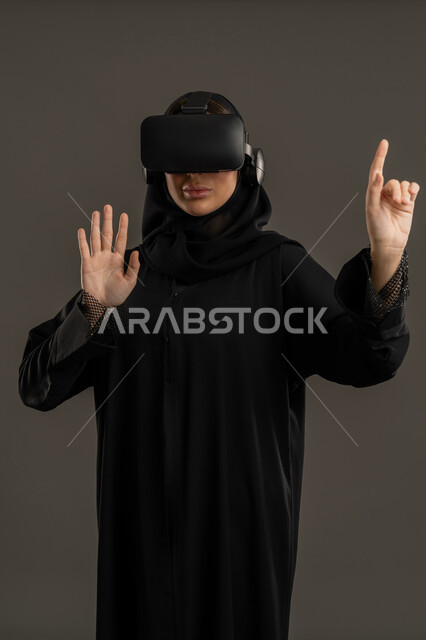 Virtual world simulation. Playing recreational video games. Using the sense of touch to touch holographic elements. Integrating fun and entertainment with the virtual world. A portrait of an Arab Gulf Emirati woman wearing a black abaya, hijab, and virtual reality (VR) glasses. A Saudi woman raising her hand to touch something. A technological device and modern technology. A black background.
