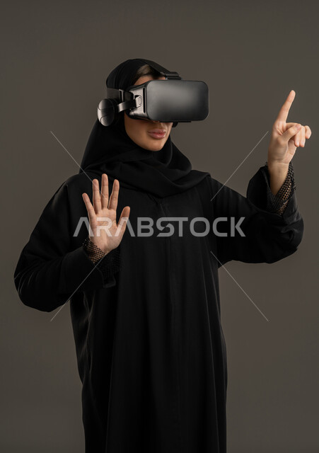 Using the sense of touch to touch holographic elements, integrating fun and entertainment with the virtual world. A portrait of an Arab Gulf Emirati woman wearing a black abaya and hijab, wearing virtual reality (VR) glasses. A Saudi woman raising her hand to touch something. A technological device and modern technology. A black background.