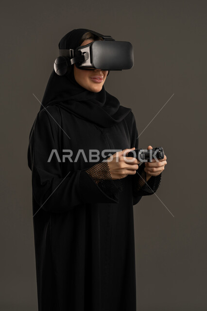 دمج المتعة والتسلية مع العالم الافتراضي، قضاء وقت فراغ ممتع ، بورتريه لامرأة عربية خليجية اماراتية ترتدي الحجاب والعباءة السوداء ونظارة الواقع الافتراضى ( في آر )، تمسك بيدها عصا التحكم تقوم باللعب في ألعاب الفيديو الترفيهية، سيدة سعودية بتعابير السرور والسعادة ، جهاز تكنولوجي وتقنية حديثة ، خلفية سوداء .