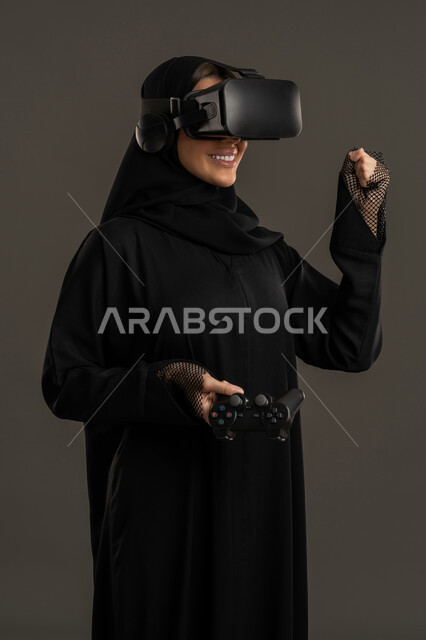 A technological device and modern technology, combining fun and entertainment with the virtual world. A portrait of an Arab Gulf Emirati woman wearing a hijab, a black abaya, and virtual reality (VR) glasses, holding a joystick while playing an entertainment video game. A Saudi woman with an expression of pleasure and happiness, spending a pleasant leisure time, black background.