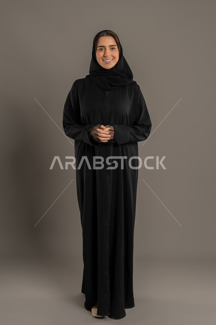 الوقوف باستقامه وايدى متشابكة ،مفهوم الأنوثة والنعومة ، الاناقة والاهتمام بالمظهر الخارجي, بورتريه لامراة عربية خليجية إماراتية ترتدي العباءة السوداء والحجاب تقف باعتدال وثقة بالنفس ، سيدة سعودية محجبة تنظر إلى الكاميرا بإيماءات السعادة والسرور , صورة الجسم بالكامل, خلفية سوداء .