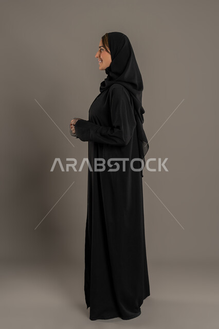 The concept of femininity and softness, gestures of self-confidence, elegance, and attention to one's appearance. A side portrait of an Emirati Gulf Arab woman wearing a black abaya and hijab, standing upright. A veiled Saudi woman looks at something with a happy expression. Full-body image, black background.