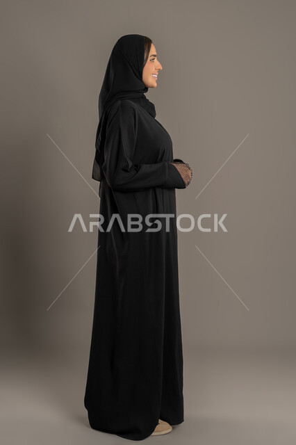 الوقوف بثقة بالنفس, مفهوم الأنوثة والنعومة, الاناقة والاهتمام بالمظهر الخارجي, بورتريه من الجانب لامراة عربية خليجية إماراتية ترتدي العباءة السوداء والحجاب تقف باستقامة ، سيدة سعودية محجبة تنظر الى شئ ما بايماءات السعادة , صورة الجسم بالكامل, خلفية سوداء .