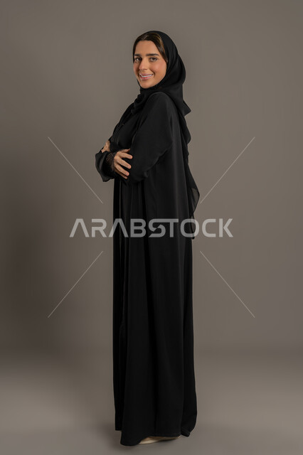 الاناقة والاهتمام بالمظهر الخارجى، بورتريه من الجانب لامراة عربية خليجية إماراتية ترتدي العباءة السوداء والحجاب تقف باستقامة وايدي متقاطعة ، سيدة سعودية محجبة تنظر الى الكاميرا بايماءات السعادة والثقة بالنفس , صورة الجسم بالكامل, خلفية سوداء .