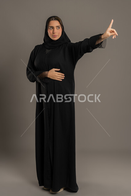 إيماءات الصمت وامتناع التدخل ، بورتريه لامراة عربية خليجية إماراتية ترتدي العباءة السوداء والحجاب ، تقف بجدية وتنظر إلى شئ ما , الهدوء والسكون وعدم الإزعاج، سيدة سعودية محجبة ترفع اصبع السبابة للنهى ، التوقف عن الكلام , صورة الجسم بالكامل, خلفية سوداء .