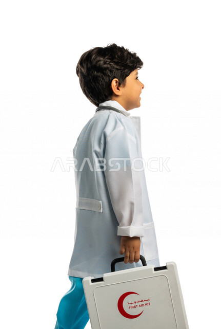 بورتريه بمنظر جانبي لفتى عربي خليجي سعودي ، يحمل بيده حقيبة الإسعافات الأولية ، يقوم بالسير بمفهوم لغة الجسد المختلفة ، يرتدي زي الطبيب ، الطبيب الصغير ، يحلم بأن يصبح طبيب ، المهنة المستقبلية ، مفهوم الطب والرعاية الصحية ، خلفية بيضاء