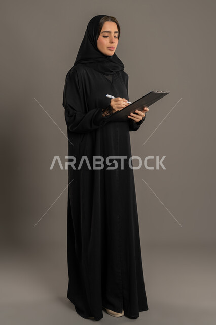 تدوين وتسجيل المعلومات على الورق، تنظيم وإدارة الأعمال, بورترية لامرأة عربية خليجية اماراتية محجبة ترتدي العباءة السوداء تمسك بيدها قلما وتقوم بكتابة الملاحظات،شابه شعودية تقوم بكتابة المهام و الاهداف اليومية، خلفية الجسم بالطول الكامل، خلفية سوداء