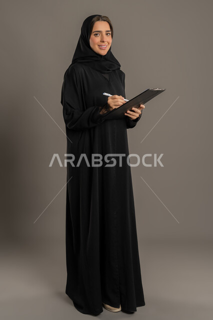 تنظيم وإدارة الأعمال, تدوين وتسجيل المعلومات على الورق, بورترية لامرأة عربية خليجية اماراتية محجبة ترتدي العباءة السوداء تمسك بيدها قلما وتقوم بكتابة الملاحظات، شابه شعودية تقوم بكتابة المهام و الاهداف اليومية، خلفية الجسم بالطول الكامل، خلفية سوداء