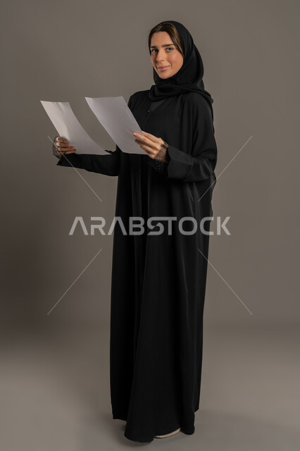 ملفات ووثائق خاصة بالعمل, متابعة المراسلات والسجلات، بورتريه  لامرأةعربية خليجية اماراتية ترتدي العباءة والحجاب مبتسمة تحمل بيديها اوراق بيضاء و تنظر اليهم، امراة سعودية تبحث عن افكار وخطط،الجسم بالطول الكامل، خلفية سوداء