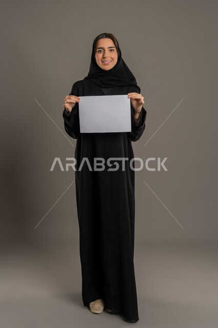 عرض و اعلان تسويقي, بورتريه لإمرأة عربية خليجية اماراتية محجبة  ترتدي العباءة السوداء تمسك بيديها لوحة بيضاء مستطيلة فارغة، شابة سعودية تنظر بايماءات السعادة والسرور، لوحات توضيحية ووسائل تعليمية, موك اب مستطيل أبيض, الجسم بالطول الكامل, خلفية سوداء