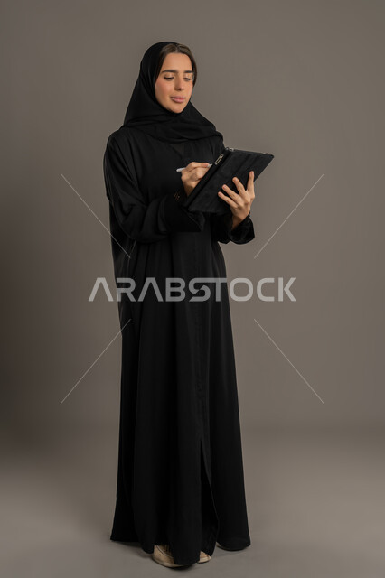 تصفح الملاحظات الهامة، متابعة المراسلات والسجلات، بورتريه لامراة عربية خليجية اماراتية محجبة ترتدي العباءة السوداء تحمل بيديها حامل اوراق تقوم بتدووين الملاحظات ، سيده سعودية تقوم بانجاز المهام المطلوبة،الجسم بالطول الكامل، خلفية سوداء