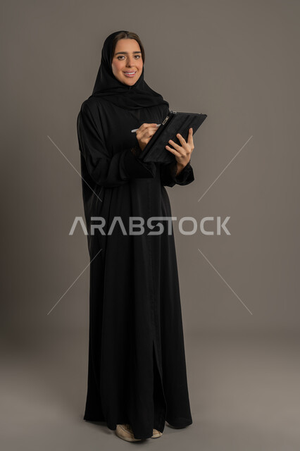التدقيق في قراءة الملاحظات، متابعة المراسلات والسجلات، بورتريه لامراة عربية خليجية اماراتية محجبة ترتدي العباءة السوداء تحمل بيديها حامل اوراق تقوم بتدووين الملاحظات ، سيده سعودية مبتسمه  تقوم بانجاز المهام المطلوبة،الجسم بالطول الكامل، خلفية سوداء