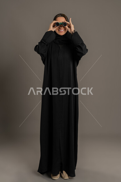 Passion, love of exploration and adventure, a Saudi woman seeing distant things through a telescope, future planning for goals and ambitions, portrait of a smiling veiled Emirati Gulf Arab woman wearing a black abaya looking through binoculars, full body, black background