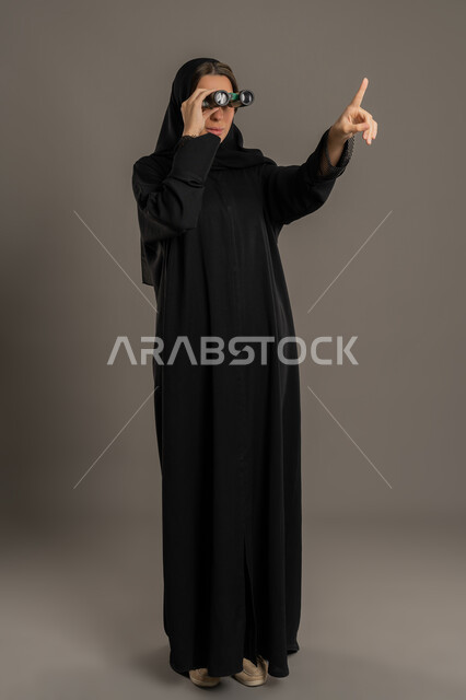 رؤية الأشياء البعيدة، التخطيط المستقبلي للأهداف والطموحات، حب الاستكشاف والمغامرة، بورتريه لشابة عربية خليجية اماراتية محجبة ترتدي العباءة تمسك منظار ثنائي العينين، امراة سعودية تقوم بالاشارة باصبع السبابة الى مكان ما , الجسم بالطول الكامل، خلفية سوداء