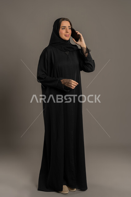 قضاء اوقات ممتعة بتبادل الاخبار والأحاديث مع الاصدقاء والأهل، بورتريه لإمرأة عربية خليجية اماراتية محجبة ترتدي العباءة السوداء تقوم بالتحدث عبر الهاتف المحمول، سيده سعوديه تقوم بإجراء مكالمة هاتفية بايماءات السعادة و السرور، الجسم بالطول الكامل، خلفية سوداء