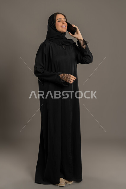التواصل مع الاهل والاصدقاء، الدردشة والتحدث عبر الجوال, بورتريه لإمرأة عربية خليجية اماراتية محجبة ترتدي العباءة السوداء تقوم بالتحدث عبر الهاتف المحمول، سيده سعوديه تقوم بإجراء مكالمة هاتفية بايماءات السعادة و السرور، الجسم بالطول الكامل، خلفية سوداء