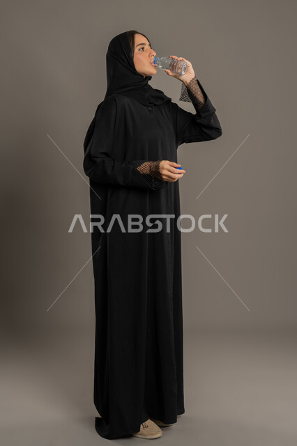المحافظة على سلامة الجهاز المناعي للجسم، سيدة سعودية تشعر بالعطش والجفاف، بورتريه لامراة عربيه خليجيه امارتية ترتدي العباءة السوداء تقوم بشرب الماء من زجاجة بلاستيكية صغيرة، المداومة على شرب المياه، الجسم بالطول الكامل، خلفية سوداء