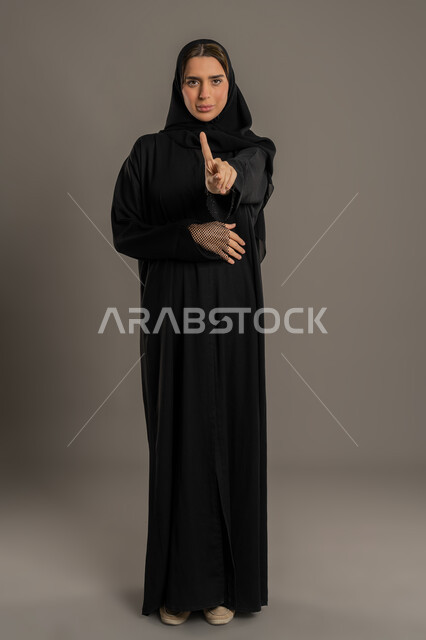 الصمت و التوقف عن الكلام، سيد سعوديه تقوم بالنهي عن فعل أمر ما، بورتريه لامراة عربية خليجيه اماراتية ترتدي العباءة السوداء وتشير باصبع السبابة بإيماءات التوقف وعدم الاقتراب، صورة الجسم بالطول الكامل، خلفية سوداء