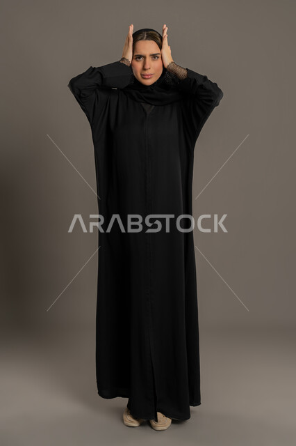التعجب والتفاجؤ من مشاهدة شيء ما،سيده سعودية تنظر الي الكاميرا بتعابير الاستغراب والاندهاش, بورتريه لامرأة عربية خليجية امارتية محجبة ترتدي عباءة سوداء ترفع يديها بإيماءات الذهول والصدمة، صورة الجسم بالطول الكامل، خلفية سوداء