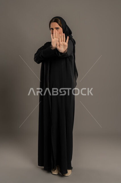 مفهوم الرفض والامتناع والانزعاج، سيد سعوديه تقوم بالنهي عن فعل أمر ما، بورتريه لامراة عربية خليجيه اماراتية ترتدي العباءة السوداء و  ترفع يدها بإيماءات التوقف وعدم الاقتراب، صورة الجسم بالطول الكامل، خلفية سوداء
