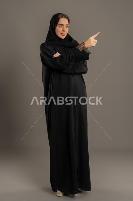 الإشارة إلى شيء محدد, امرأةعربية خليجية اماراتية تنظر الى شيء ما بتعابير البشاشة والسرور, بورتريه لسيدة سعودية مبتسمة ترتدي العباءة السوداء ترفع إصبع السبابة إلى الاعلى بتعابير التأكد والتحديد, الجسم بالطول الكامل, خلفية سوداء