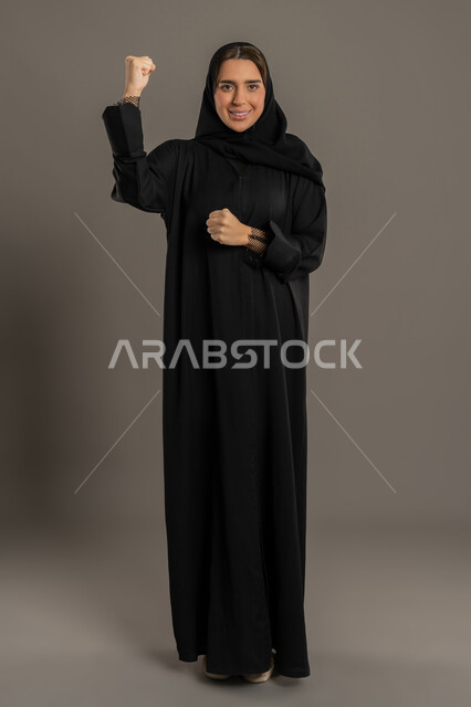 مفهوم الانجاز وتحقيق الاهداف، الانتصار والفوز والنجاح، بورتريه لامراة عربيه خليجية اماراتية ترتدي العباءة السوداء تنظرالي الكاميرا و  ترفع قبضة يديها للاعلى بإيماءات الفرح والسرور, سيدع سعوديه تشعربالسعادة والقوة، صورة الجسم بالطول الكامل, خلفية سوداء