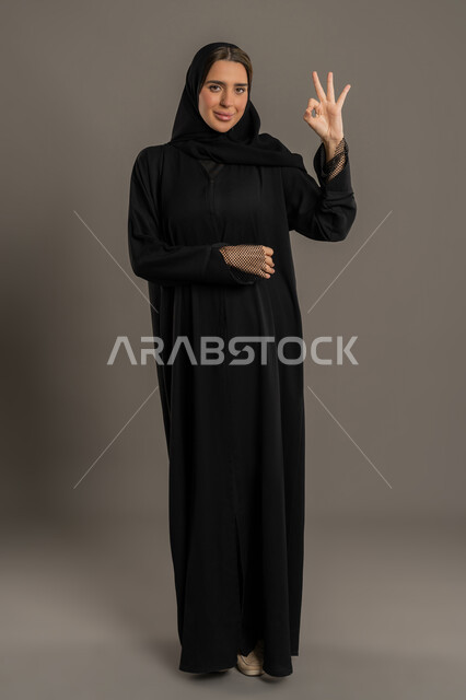 النظر الى الكاميرا بتعابير السرور، سيدة سعودية تشعر بالإعجاب، بورتريه لامراة عربية خليجية اماراتية مبتسمة محجبة ترتدي العباءة السوداء ترفع اصبع الابهام للاعلى بإيماءات علامة الجودة و الامتياز, الشعور بالرضى و القبول و الموافقة ,الجسم بالطول الكامل,خلفية سوداء