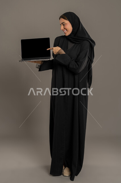 Using modern and advanced technological devices, managing and organizing business affairs, interacting with technology and providing services, portrait of a veiled Emirati Arab woman wearing a black abaya, a blank black screen on a laptop, black background