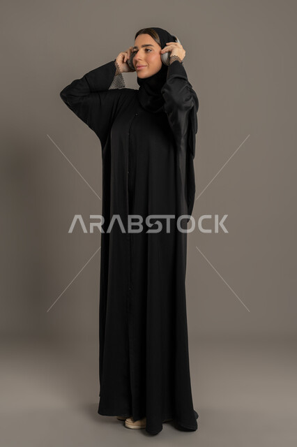 Enjoying listening to your favorite music, modern technology devices, a Saudi woman having a good time listening to songs, a portrait of an Arab Gulf Emirati woman wearing a hijab and abaya, putting on headphones, full body, black background