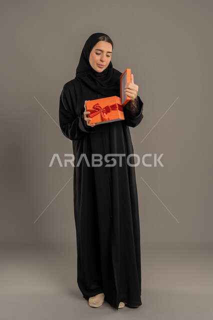التعبير عن المحبة والتقدير,الاحتفال بأعياد الميلاد, تقديم الهدايا في المناسبات السعيدة, شابة سعودية تفتح صندوق هدايا بإيماءات السرور, بورتريه لامرأة عربية خليجية اماراتية محجبة ترتدي العباءة تمسك بيديها علبة هداية, الجسم بالطول الكامل، خلفية سوداء