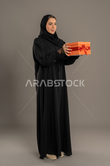 تقديم الهدايا في المناسبات السعيدة, الاحتفال بأعياد الميلاد, التعبير عن المحبة والتقدير, بورتريه لامرأة عربية خليجية اماراتية محجبة ترتدي العباءة تمسك بيديها علبة هداية, شابة سعودية تنظر إلى الكاميرا بإيماءات السعادة والسرور, الجسم بالطول الكامل، خلفية سوداء