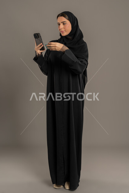 قضاء وقت ممتع عبر الجوال مع الأصحاب, التكنولوجيا والتقنية الحديثة، تصفح مواقع التواصل الاجتماعي، بورتريه لامرأة عربية خليجية اماراتية محجبة ترتدي العباءة تقوم بإرسال الرسائل النصية عبر الهاتف المحمول،الجسم بالطول الكامل, خلفية سوداء